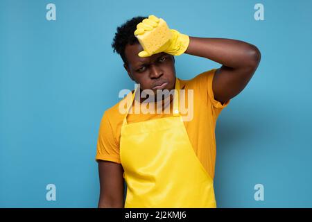 Jeune africain fatigué portant des gants en caoutchouc jaune pour la protection des mains essuyant la sueur tout en nettoyant la maison isolée sur fond bleu Banque D'Images