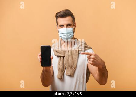 Portrait d'un homme brunette avec masque médical en t-shirt blanc pointant vers un téléphone portable et souriant à l'appareil photo, recommandant un gadget ou une application mobile. Studio d'intérieur isolé sur fond orange Banque D'Images