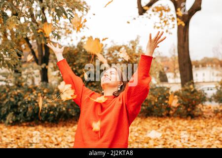 Jolie femme gaie et souriante qui jette l'érable feuilles d'or parc naturel. Porter un chandail rouge. Week-end, vacances Banque D'Images