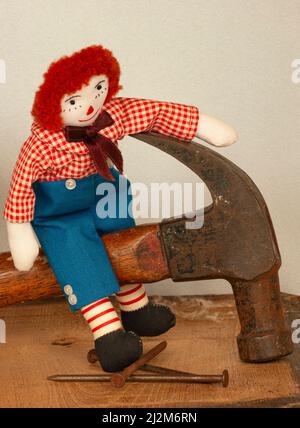 Andy raggedy est assis sur la poignée d'un marteau avec des clous et du bois dans un cadre d'atelier. Le marteau est grand. Andy est une poupée de chiffon de petit garçon avec des cheveux rouges. Banque D'Images