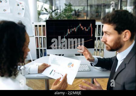 Des collègues multiraciaux à succès, des traders de crypto-monnaies, une femme afro-américaine et un homme caucasien, s'assoient à une table dans un bureau moderne, travaillent avec des diagrammes de stock et des documents, analysent la dynamique Banque D'Images