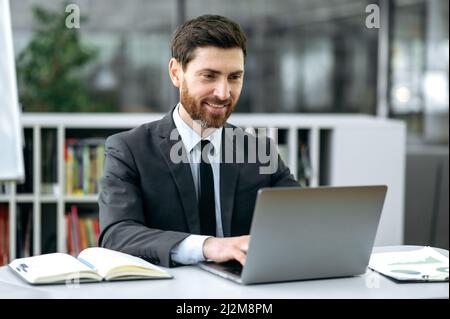 Beau homme de race blanche, directeur, avocat, économiste, assis à son bureau devant un ordinateur portable, dans un bureau moderne, dans des vêtements d'affaires élégants, effectue la correspondance de travail avec un client ou un employé, sourit Banque D'Images