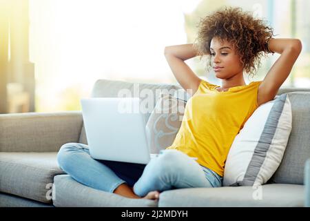 Diffuser ses émissions préférées ce week-end. Prise de vue d'une jeune femme utilisant un ordinateur portable lors d'une journée de détente à la maison. Banque D'Images