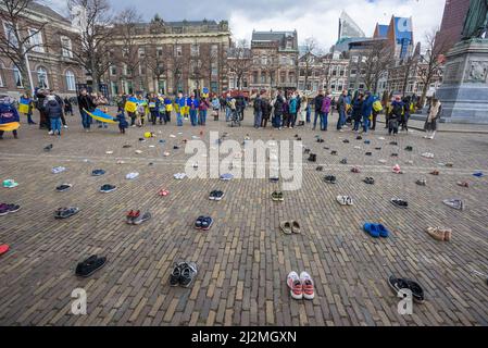 Des chaussures disposées en rangées sur la plaine de Het, tandis que des adultes et des enfants drapaient dans les drapeaux ukrainiens et tiennent des écriteaux lors de la manifestation de l'installation artistique ukrainienne. Une installation d'art par des membres de la communauté ukrainienne aux pays-Bas a été installée pour attirer l'attention, en particulier sur le gouvernement. L'installation avait des chaussures disposées en rangées dans la plaine de Het (Plain Square) dans le centre-ville de la Haye, pour attirer l'attention sur le massacre aveugle de civils et d'enfants en Ukraine, alors que l'invasion russe de l'Ukraine entrait dans les sixième semaines. Banque D'Images