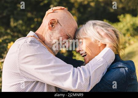 Un couple de personnes âgées affectueux se touchant la tête tout en se tenant debout à l'extérieur. Couple senior heureux partageant un moment romantique dans un parc. Joyeux el Banque D'Images
