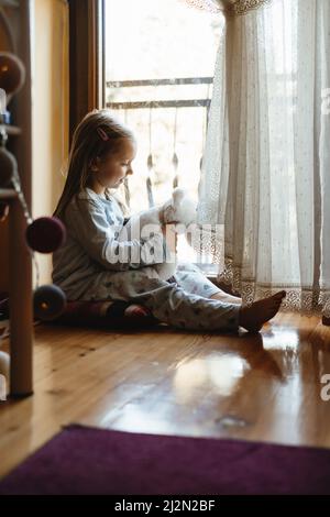 Enfant assis sur le sol et jouant à la maison. Ambiance rêveuse et ensoleillée. Fille regardant par la fenêtre de l'arrière. Un adorable enfant qui attend pour une promenade Banque D'Images