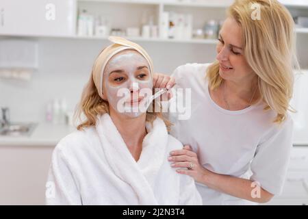 Cosmétologie. Une esthéticienne professionnelle en masque médical et gants en caoutchouc applique un masque au visage du client. Le concept de l'aide professionnelle dans les salons de beauté pendant une pandémie. Banque D'Images