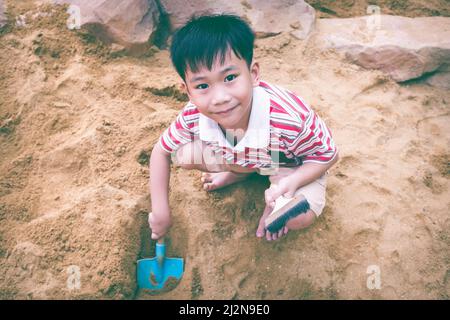 Vue de dessus. Enfant heureux jouant avec le sable et souriant. Adorable garçon asiatique a le plaisir de creuser dans le sable un jour d'été et de regarder la caméra. Tonne vintage Banque D'Images