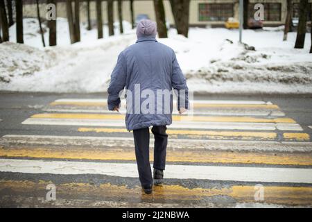 Femme traverse la route sur zébra. Le pensionné en Russie descend la rue. L'homme marche sur le passage piéton. Prise de vue depuis l'arrière. Banque D'Images