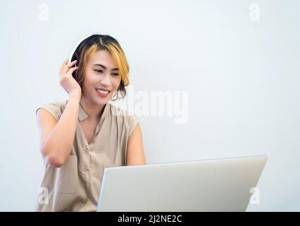 Femme asiatique avec cheveux courts tendance utilisant des écouteurs blancs tout en travaillant avec un ordinateur portable, freelance regardant l'écran sur fond blanc de mur Banque D'Images