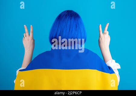 Soutenir le concept de l'Ukraine. Femme avec perruque bleue à cheveux courts, blouse blanche et drapeau national de l'Ukraine sur ses épaules debout avec ses mains vers le haut avec v or Banque D'Images