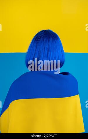 Soutenir le concept de l'Ukraine. Femme avec perruque bleue à cheveux courts, blouse blanche et drapeau national de l'Ukraine sur ses épaules debout avec affiche blanche jaune dans Banque D'Images