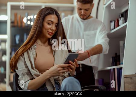 Une femme heureuse utilisant un smartphone tout en se coiffant au salon de beauté Banque D'Images