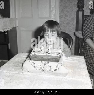 1961, historique, à la maison, une petite fille assise à une table, soufflant les bougies sur son gâteau d'anniversaire, Stockport, Manchester, Angleterre, Royaume-Uni. Banque D'Images