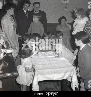 1961, historique, à la maison avec des parents et grand-père avec son pipe, une petite fille, aidée par d'autres petits enfants, soufflant les bougies sur son gâteau d'anniversaire, Stockport, Manchester, Angleterre, Royaume-Uni. Banque D'Images