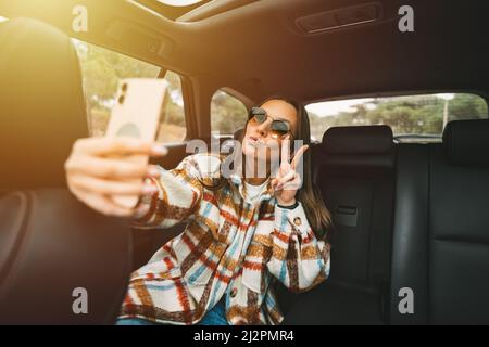 Jeune femme élégante assise sur le siège arrière dans la voiture et faisant selfie avec un smartphone. Femme prenant des photos avec le téléphone en étant assise dans un taxi Banque D'Images