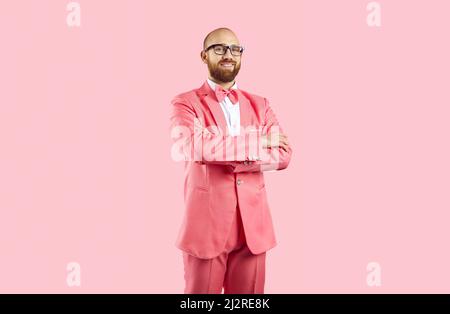 Joyeux homme confiant dans un costume rose et des lunettes debout avec les bras repliés sur fond rose Banque D'Images