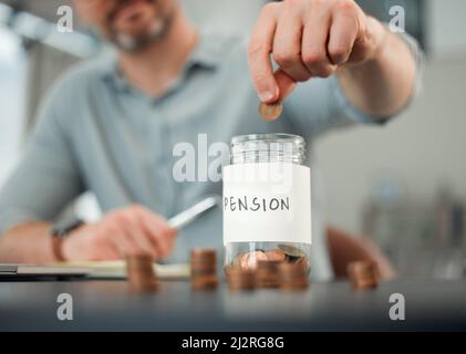 Il est important d'économiser. Un petit coup de feu d'un homme méconnaissable mettant des pièces dans un pot marqué PENSION tout en travaillant sur ses finances à un bureau à la maison. Banque D'Images