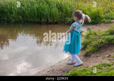 Une fille dans une robe bleue jette des cailloux dans l'eau. Un enfant joue avec la rivière dans la nature. Un bébé habillé avec élégance s'amuse près du lac. CHIL Banque D'Images