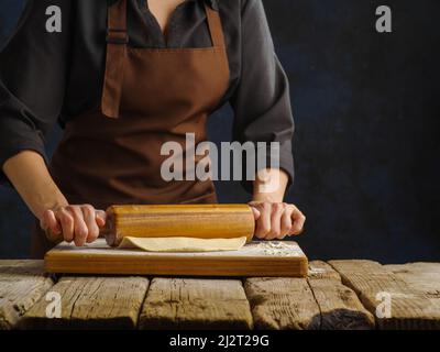 Préparation de pâte par les mains d'un chef professionnel sur une planche à découper, fond sombre. Le chef roule la pâte avec un rouleau en bois. Banque D'Images