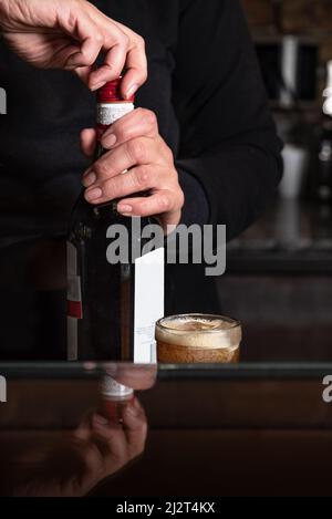 Fermer la bouteille en verre de boisson alcoolisée avec la main droite. Verre servi avec de la mousse. Réflexion sur la table. Banque D'Images