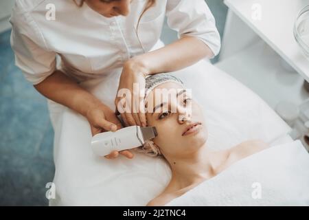 Vue de dessus des mains d'un cosmétologue faisant un nettoyage du visage par ultrasons pour une femme de race blanche adulte. Soin du visage. Machine de nettoyage Banque D'Images