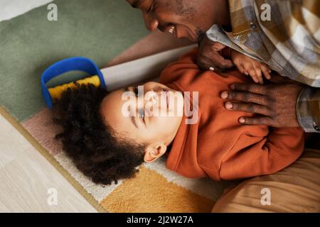 Portrait en grand angle d'un adorable garçon afro-américain jouant avec son père sur le sol dans la chambre des enfants et souriant avec joie Banque D'Images