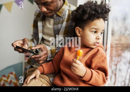 Portrait d'un petit garçon mignon tenant une lollipop et regardant la fenêtre avec le père en arrière-plan, espace de copie Banque D'Images