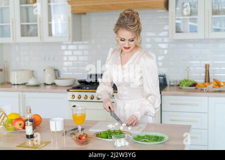 Une fille coupe une salade de fruits avec des fraises sur une planche à découper avec un couteau. Concept de cuisine en tranches de fruits frais belle femme avec maquillage et coiffure dans la robe légère aime le matin et la préparation du petit déjeuner Banque D'Images