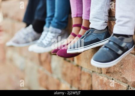 Des petits pieds qui vont dans de grands endroits. Photo courte d'enfants d'école élémentaire méconnus assis sur un mur de briques à l'extérieur. Banque D'Images