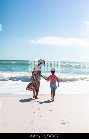 Mère afro-américaine tenant la main de la fille tout en marchant vers la mer à la plage le jour ensoleillé Banque D'Images