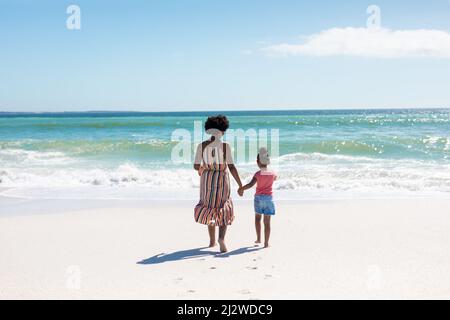 Vue arrière de la mère et de la fille afro-américaines tenant les mains tout en marchant vers la mer à la plage Banque D'Images