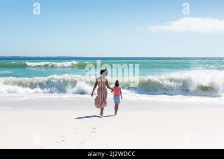 Mère et fille afro-américaines tenant les mains tout en marchant vers les vagues à la plage Banque D'Images