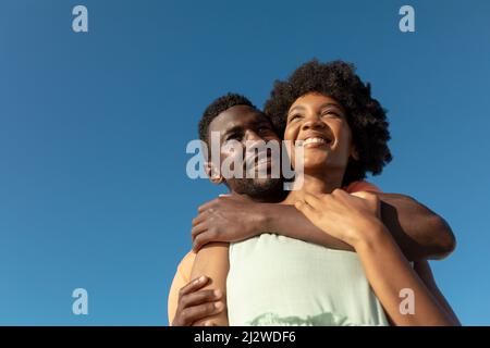 Vue en contre-angle d'un homme afro-américain souriant qui embrasse sa petite amie de derrière contre le ciel bleu Banque D'Images