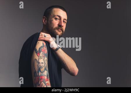 Fier homme barbu caucasien regardant directement dans l'appareil photo roulant la manche de son t-shirt pour montrer des tatouages audacieux. Photo de haute qualité Banque D'Images