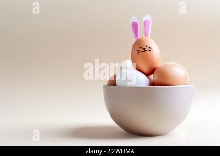 Œuf de Pâques avec un museau et des oreilles de lapin. Différents œufs dans un bol sur un fond clair avec espace de copie. Banque D'Images