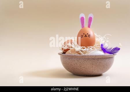 Œufs de Pâques et œuf de lapin avec oreilles de lapin et museau dans un bol de foin sur fond pastel avec espace de copie. Composition de Pâques. Banque D'Images