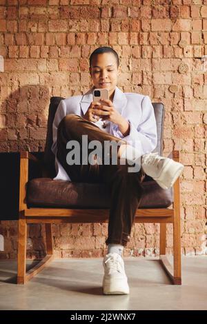 Portrait d'une femme LGBTQ multiraciale confiante assise sur une chaise rétro et utilisant un smartphone devant un mur de briques exposé Banque D'Images