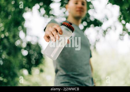 Gros plan de jeune homme coureur à capuche tenant une bouteille d'eau tout en faisant de l'entraînement dans le parc vert ensoleillé d'été. Détente après l'entraînement sportif. Banque D'Images