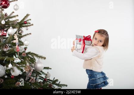 fille de 3 ans tenant une boîte cadeau enveloppée de papier argenté et de ruban rouge. Photographie de l'esprit des fêtes sur fond blanc. Arbre de Noël Banque D'Images