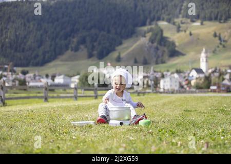 Petite fille de camping chef cuisinier déjeuner sur une cuisinière à gaz assis sur un terrain vert avec un village alpin suisse comme toile de fond au loin Banque D'Images