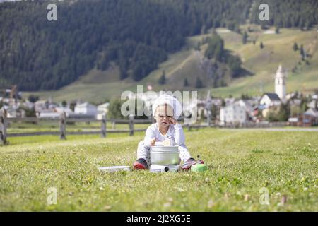 Petite fille de camping chef cuisinier déjeuner sur une cuisinière à gaz assis sur un terrain vert avec un village alpin suisse comme toile de fond au loin Banque D'Images