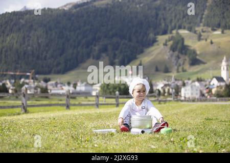 Petite fille de camping chef cuisinier déjeuner sur une cuisinière à gaz assis sur un terrain vert avec un village alpin suisse comme toile de fond au loin Banque D'Images