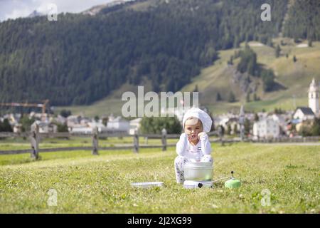 Petite fille de camping chef cuisinier déjeuner sur une cuisinière à gaz assis sur un terrain vert avec un village alpin suisse comme toile de fond au loin Banque D'Images