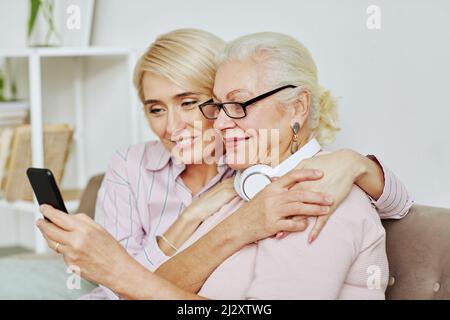 Portrait d'une fille adulte souriante qui embrasse une mère âgée et regarde l'écran d'un smartphone dans une maison confortable Banque D'Images
