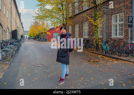 Une jeune fille étudiante souriante dans des vêtements chauds avec une coupe de papier avec café à la main se tient dans une petite rue de la vieille ville, au milieu de la brique et du woo Banque D'Images