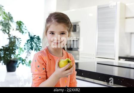 La petite fille caucasienne mange une grosse pomme rouge dans la cuisine. Belle petite fille mord et mange une pomme. Regarde l'appareil photo qui tient ses fruits. Gros plan Banque D'Images