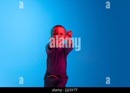 Portrait de petit pleurs garçon, enfant, enfant d'âge préscolaire est isolé capricieux sur le fond bleu de studio en lumière rouge néon. Concept des émotions de l'enfant Banque D'Images