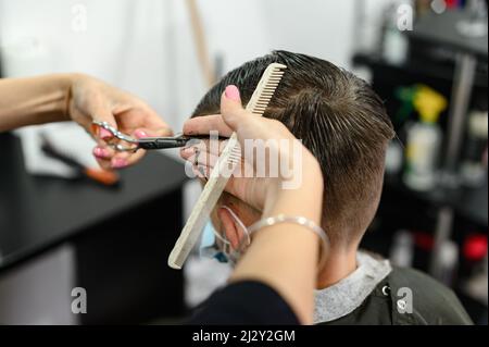 Un adolescent dans un salon de beauté obtient une coupe de cheveux, un coiffeur coupe les cheveux d'un garçon adolescent, une coupe de cheveux avec des ciseaux et un peigne. Banque D'Images