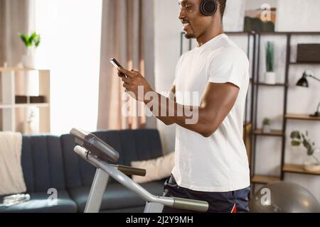 Image rognée d'un homme africain qui court sur un tapis roulant avec un téléphone dans la main et un casque dans un appartement moderne lumineux. Concept de mode de vie sain. Youn Banque D'Images